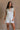 Front view of female model wearing the Kassidy White Denim Sleeveless Romper which features White Cotton Fabric, Two Front Pockets, Two Back Pockets, Fray Hem, Adjustable Tie Straps and Sleeveless