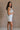 Full body side view of female model wearing the Kassidy White Denim Sleeveless Romper which features White Cotton Fabric, Two Front Pockets, Two Back Pockets, Fray Hem, Adjustable Tie Straps and Sleeveless