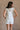 Full body back view of female model wearing the Kassidy White Denim Sleeveless Romper which features White Cotton Fabric, Two Front Pockets, Two Back Pockets, Fray Hem, Adjustable Tie Straps and Sleeveless