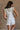 Back view of female model wearing the Kassidy White Denim Sleeveless Romper which features White Cotton Fabric, Two Front Pockets, Two Back Pockets, Fray Hem, Adjustable Tie Straps and Sleeveless