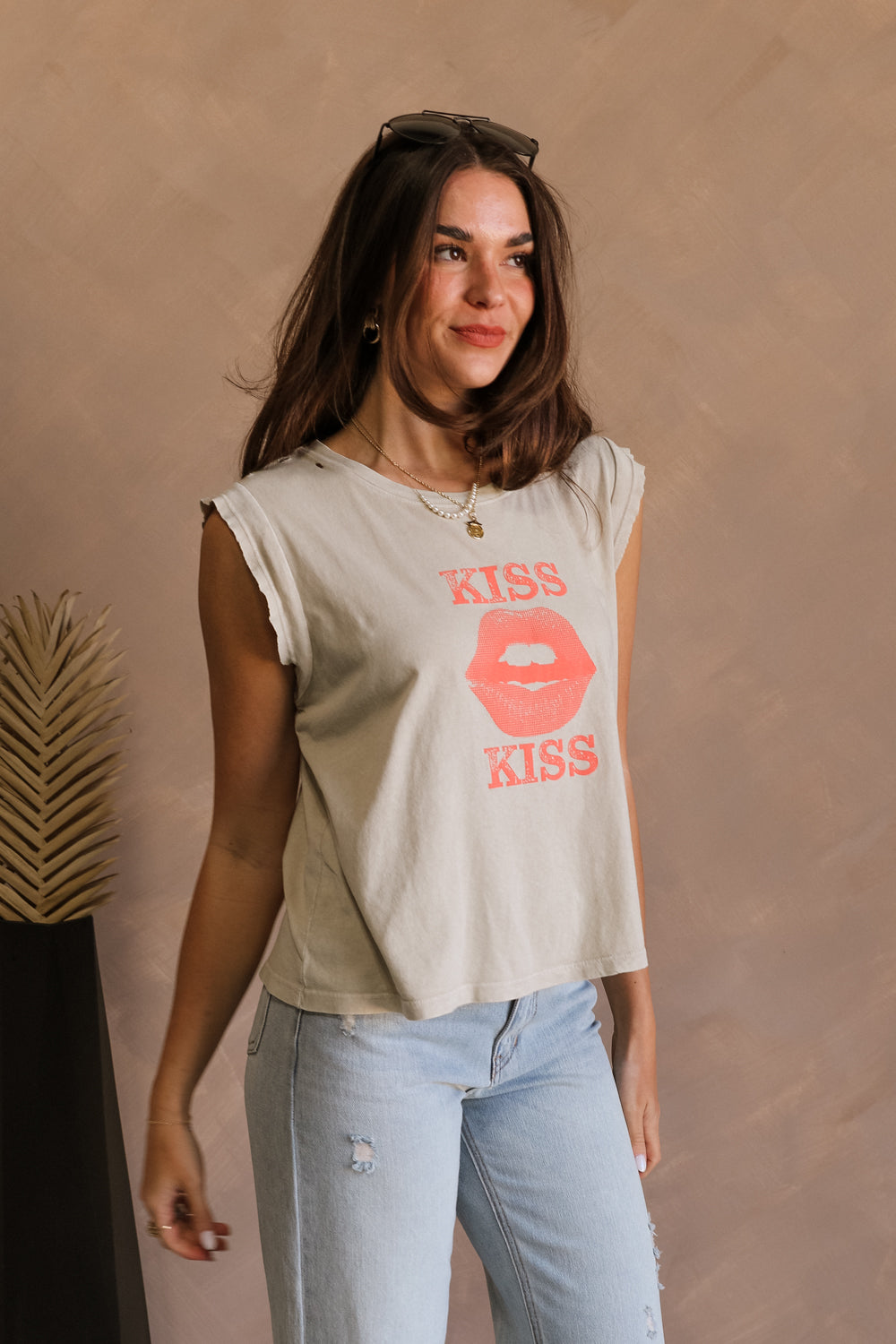 Side view of female model wearing the Kiss Stone Taupe Graphic Top which features Stone Taupe Cotton Fabric, Distressed Details, Sleeveless, Round Neckline and Kiss Graphic with Lips