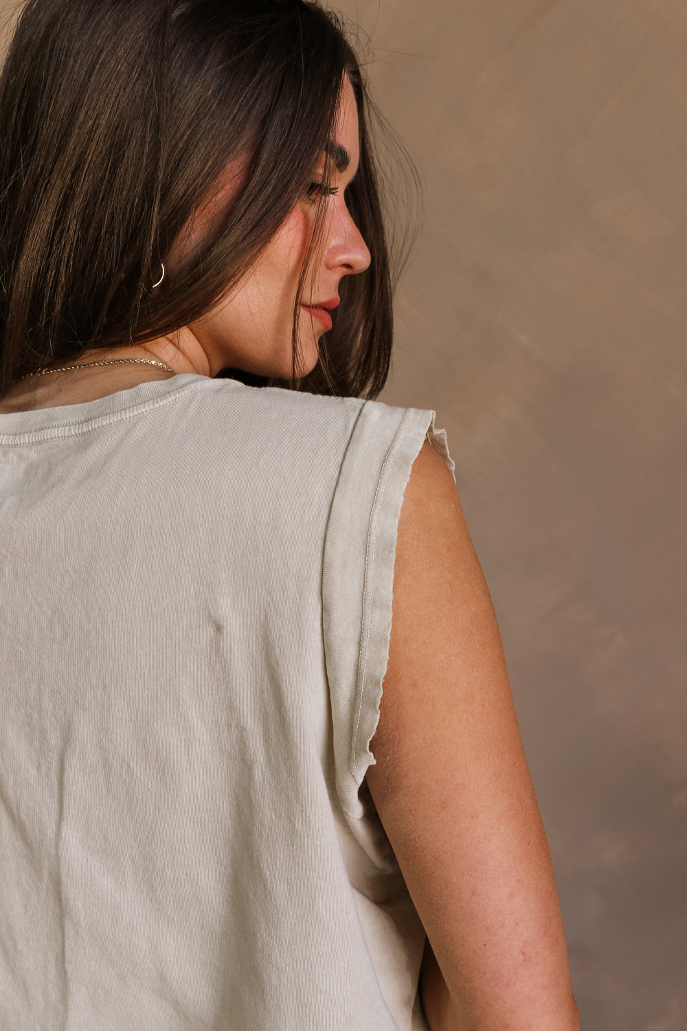 Close up back view of female model wearing the Kiss Stone Taupe Graphic Top which features Stone Taupe Cotton Fabric, Distressed Details, Sleeveless, Round Neckline and Kiss Graphic with Lips