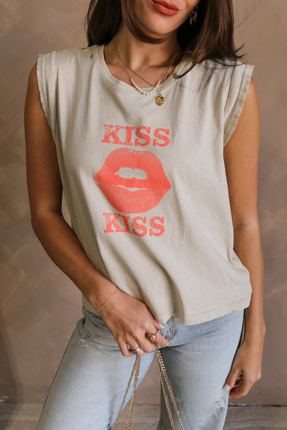 Close up view of female model wearing the Kiss Stone Taupe Graphic Top which features Stone Taupe Cotton Fabric, Distressed Details, Sleeveless, Round Neckline and Kiss Graphic with Lips