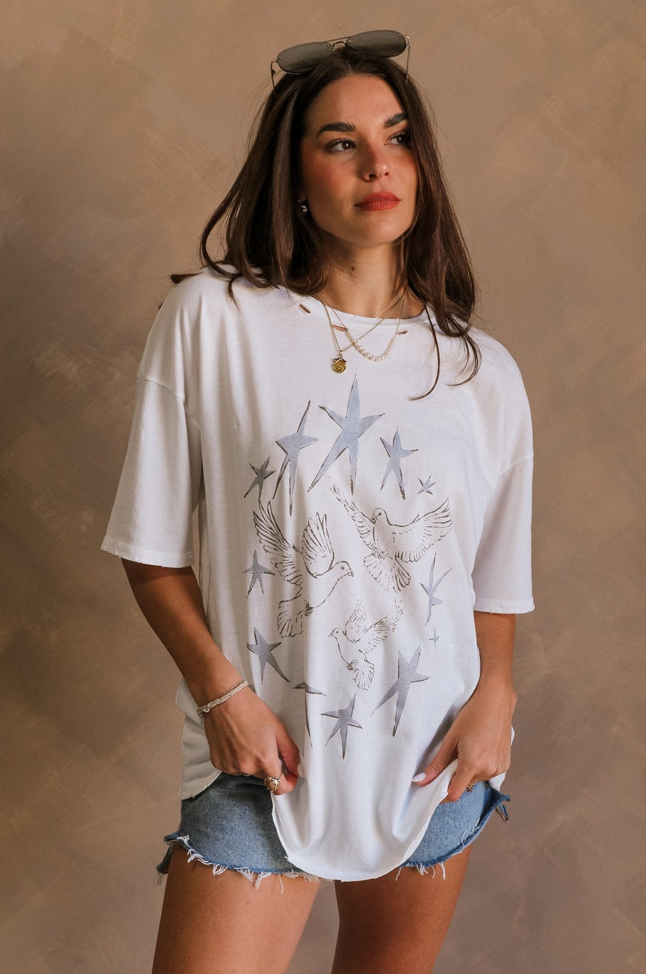 Front view of female model wearing the Stars and Birds White Distressed Graphic Top which features White Cotton Fabric, Distressed Details, Short Sleeves, Round Neckline and Stars and Birds Graphic