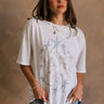 Front view of female model wearing the Stars and Birds White Distressed Graphic Top which features White Cotton Fabric, Distressed Details, Short Sleeves, Round Neckline and Stars and Birds Graphic