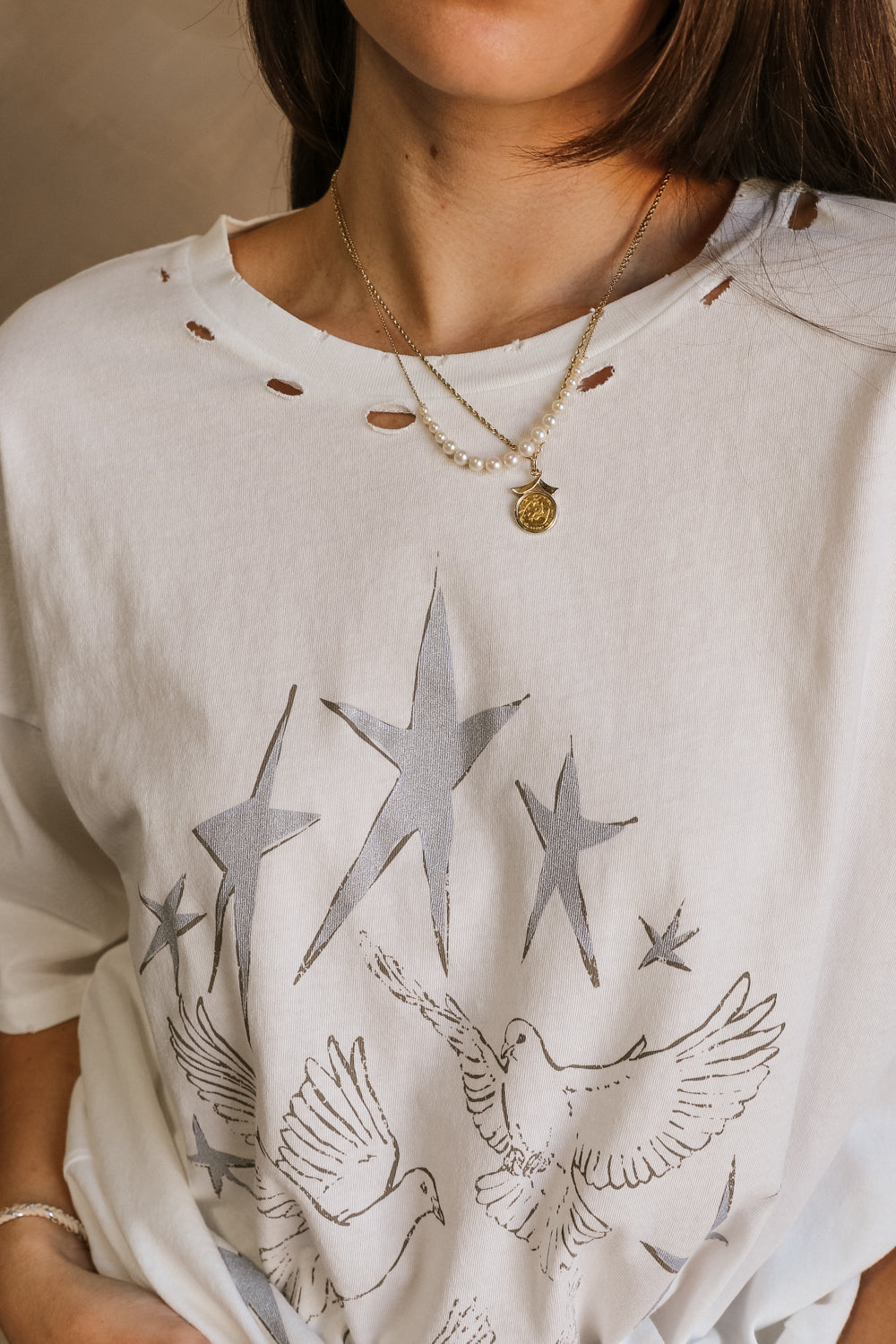 Close up view of female model wearing the Stars and Birds White Distressed Graphic Top which features White Cotton Fabric, Distressed Details, Short Sleeves, Round Neckline and Stars and Birds Graphic