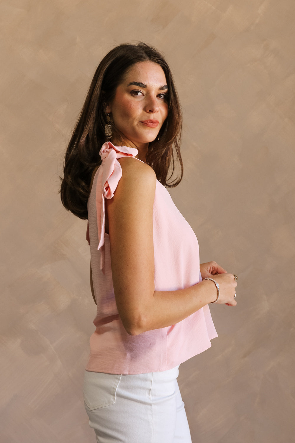 Side view of female model wearing the Jenna Blush Tie Strap Tank Top that has lightweight blush fabric, tie straps, and a v neck. Worn with white pants.