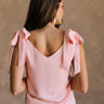 Back view of female model wearing the Jenna Blush Tie Strap Tank Top that has lightweight blush fabric, tie straps, and a v neck. Worn with white pants.