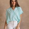 Upper body front view of female model wearing the Amalia Collared Short Sleeve Top in mint, which has short sleeves and a collar neckline. Paired with white capris. Model is holding white purse. Top is front tucked