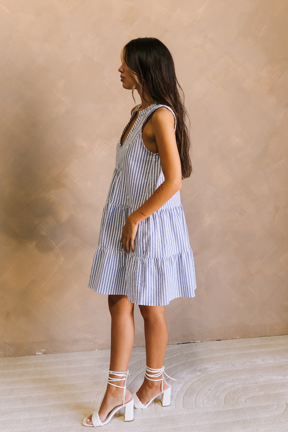 full body side view of female model wearing the Darya Blue & White Striped Mini Dress that has blue and white stripes, a notched neckline, and tiered mini skirt. Worn with white heels.