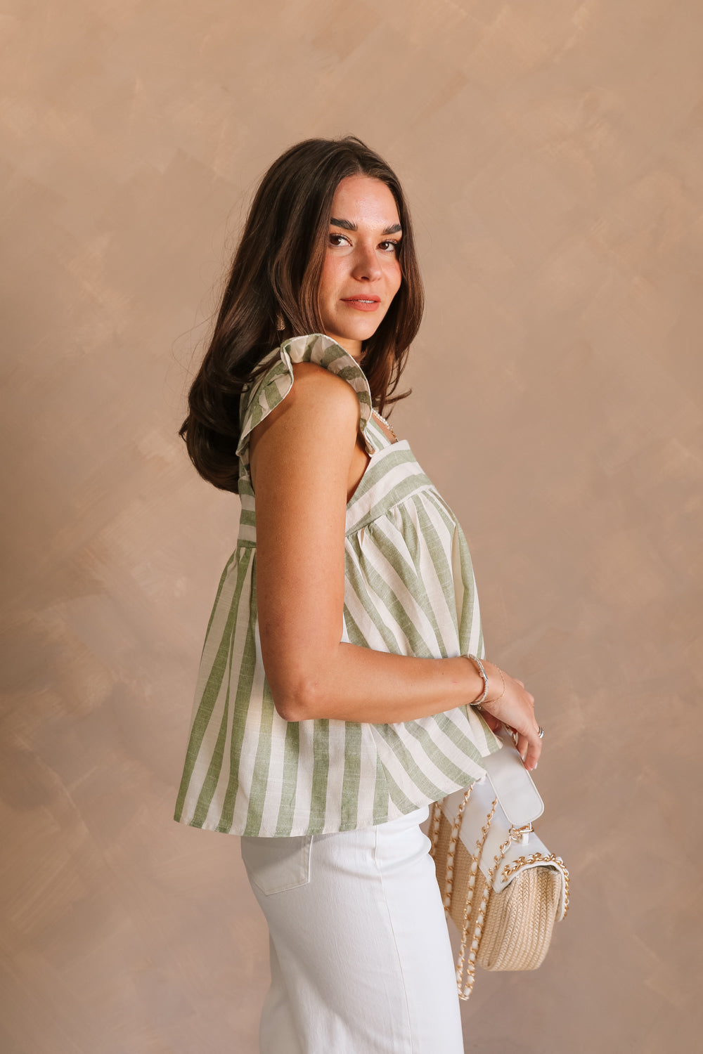 Side view of female model wearing the Coraline that has green and ivory stripes, ruffled straps, and a cropped babydoll fit. Worn with white jeans.