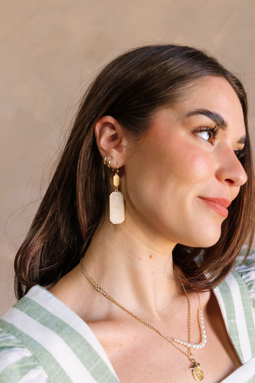 Side of female model's face; model is wearing the Addison Gold & Pearl Dangle Earrings that have a gold hexagon shape attached to gold hooks, with a gold hexagonal dangle with pearls on top.