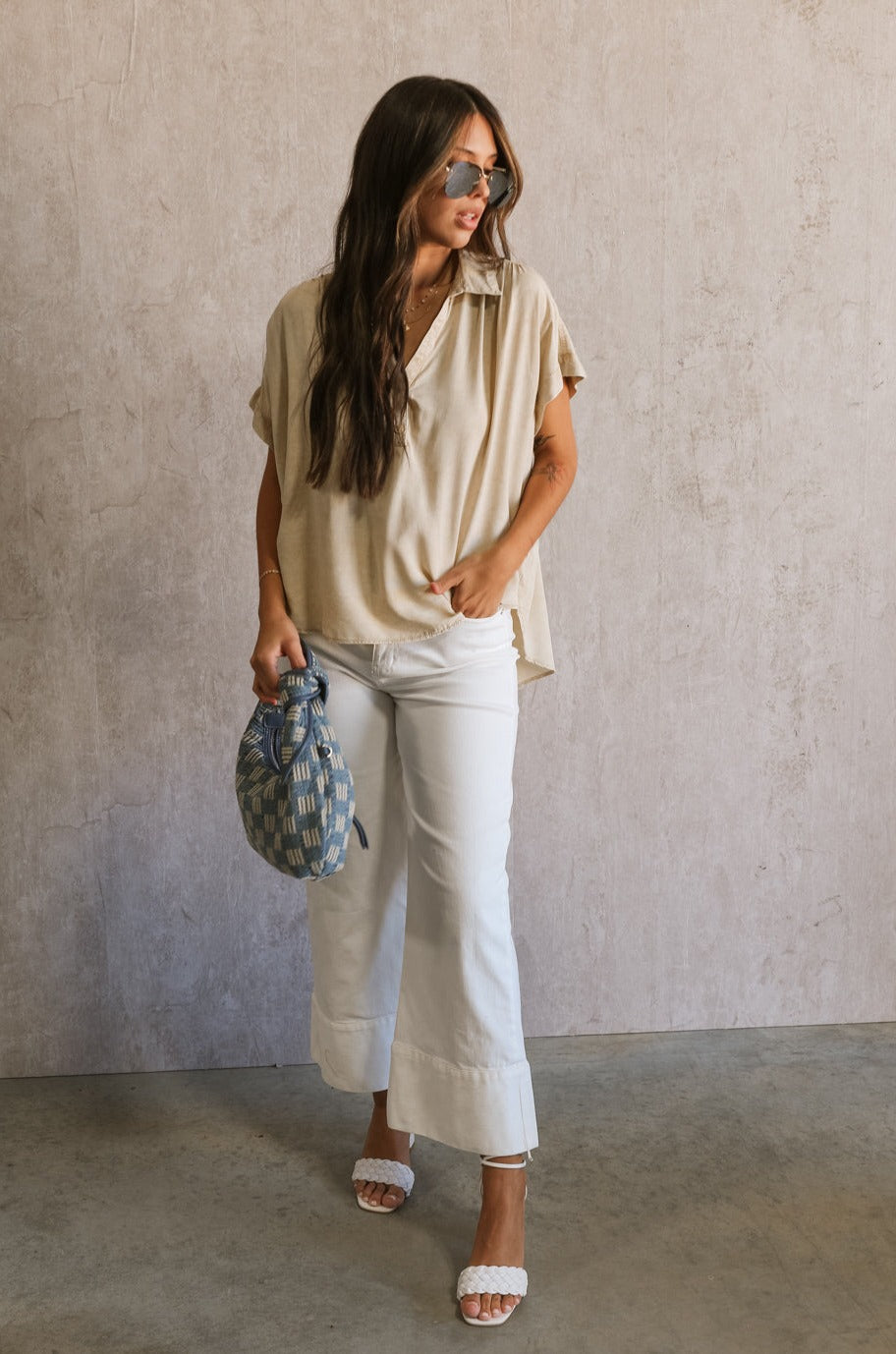 Full body front view of female model wearing the Amalia Collared Short Sleeve Top in taupe, which has short sleeves and a collar neckline. Paired with white capris. Model is holding blue purse.