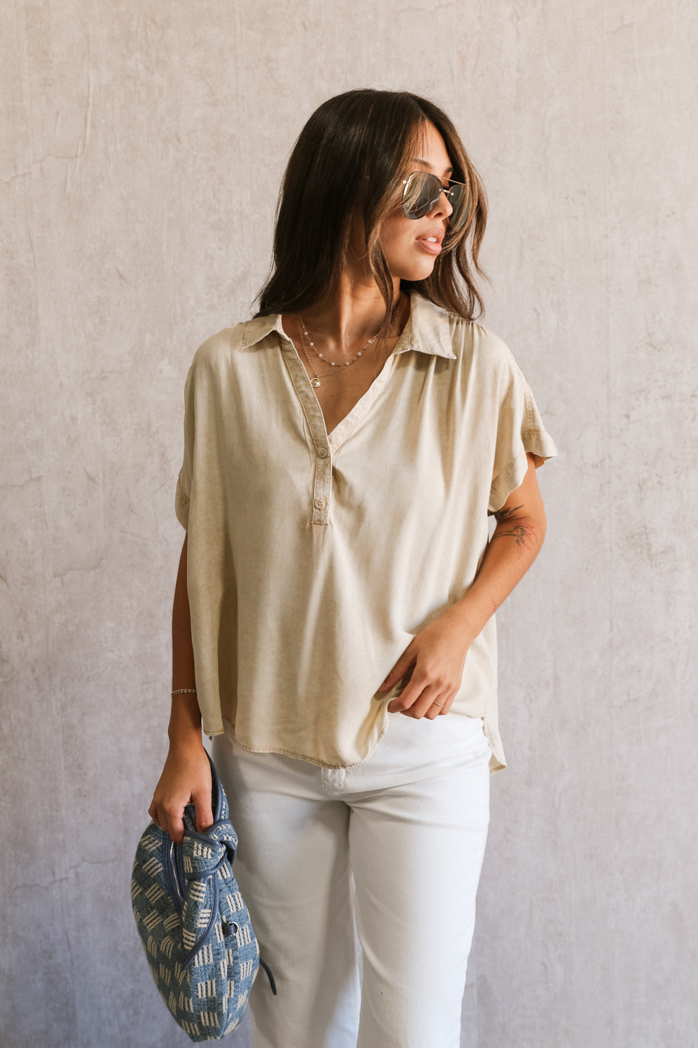 Upper body front view of female model wearing the Amalia Collared Short Sleeve Top in taupe, which has short sleeves and a collar neckline. Paired with white capris. Model is holding blue purse.