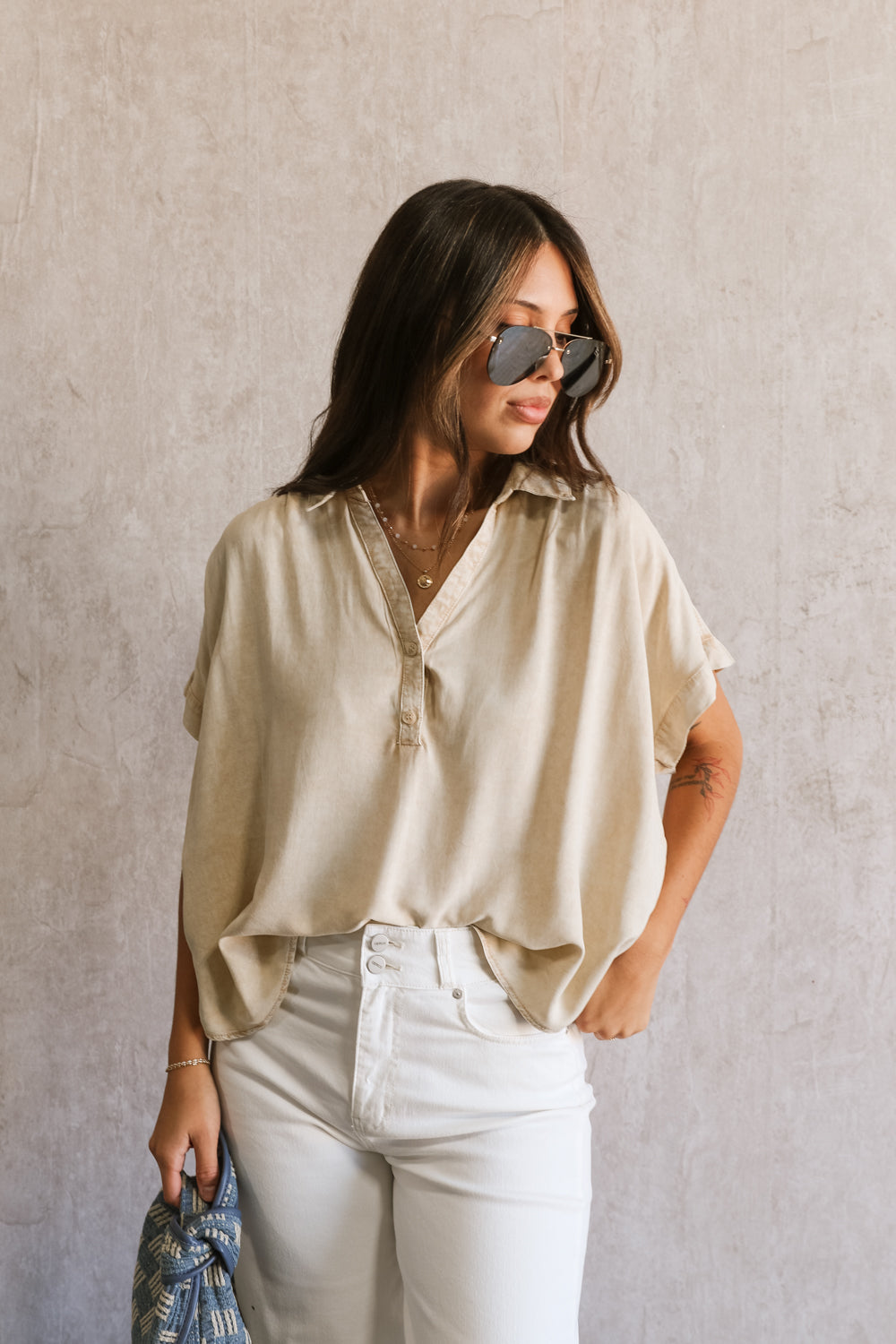 front view of female model wearing the Amalia Collared Short Sleeve Top in taupe, which has short sleeves and a collar neckline. Paired with white capris. Model is holding blue purse. Top is front tucked