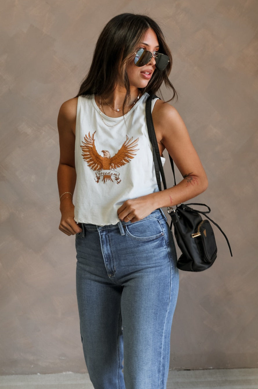 Front view of female model wearing the Free Bird Smoke Cropped Tank which features Cream Cotton Fabric, Cropped Lettuce Hem, Round Neckline, Sleeveless and Eagle Graphic