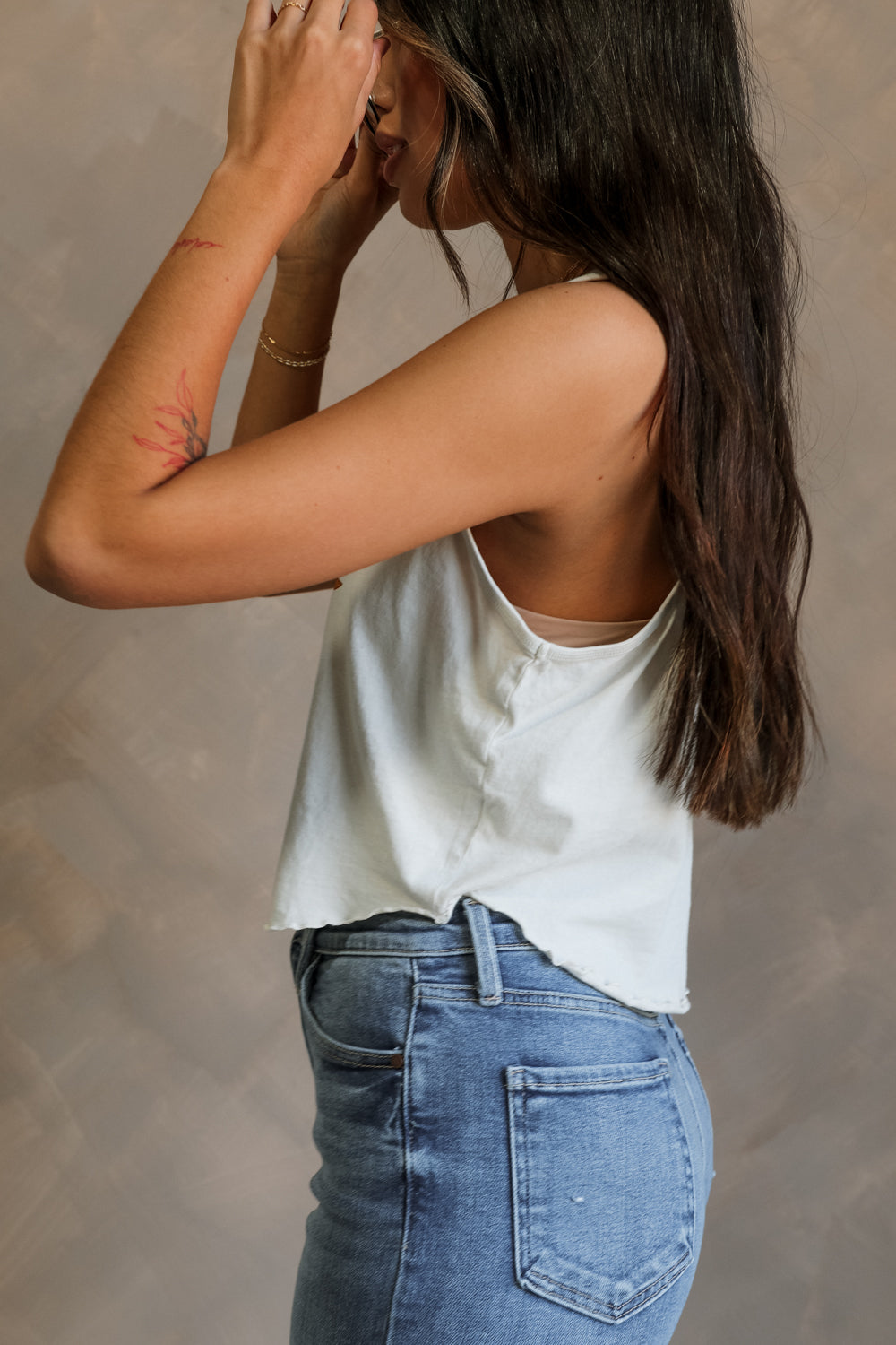 Side view of female model wearing the Free Bird Smoke Cropped Tank which features Cream Cotton Fabric, Cropped Lettuce Hem, Round Neckline, Sleeveless and Eagle Graphic
