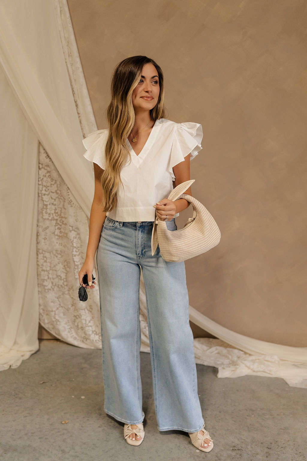 Full body front view of female model wearing the Jemma White Ruffle Top that has white fabric, short ruffled cap sleeves, a v neckline, and keyhole back. Worn with blue jeans.