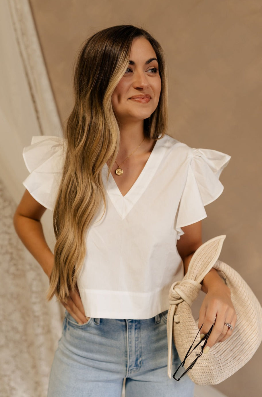 Front view of female model wearing the Jemma White Ruffle Top that has white fabric, short ruffled cap sleeves, a v neckline, and keyhole back. Worn with blue jeans.