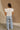 Full body back view of female model wearing the Jemma White Ruffle Top that has white fabric, short ruffled cap sleeves, a v neckline, and keyhole back. Worn with blue jeans.