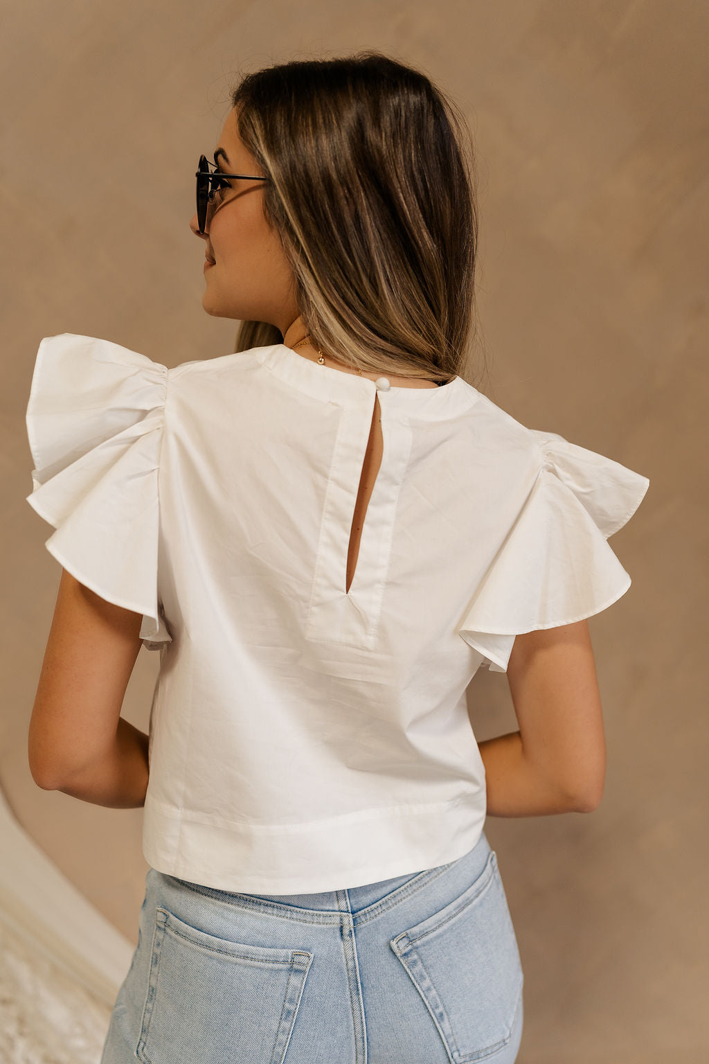Back view of female model wearing the Jemma White Ruffle Top that has white fabric, short ruffled cap sleeves, a v neckline, and keyhole back. Worn with blue jeans.