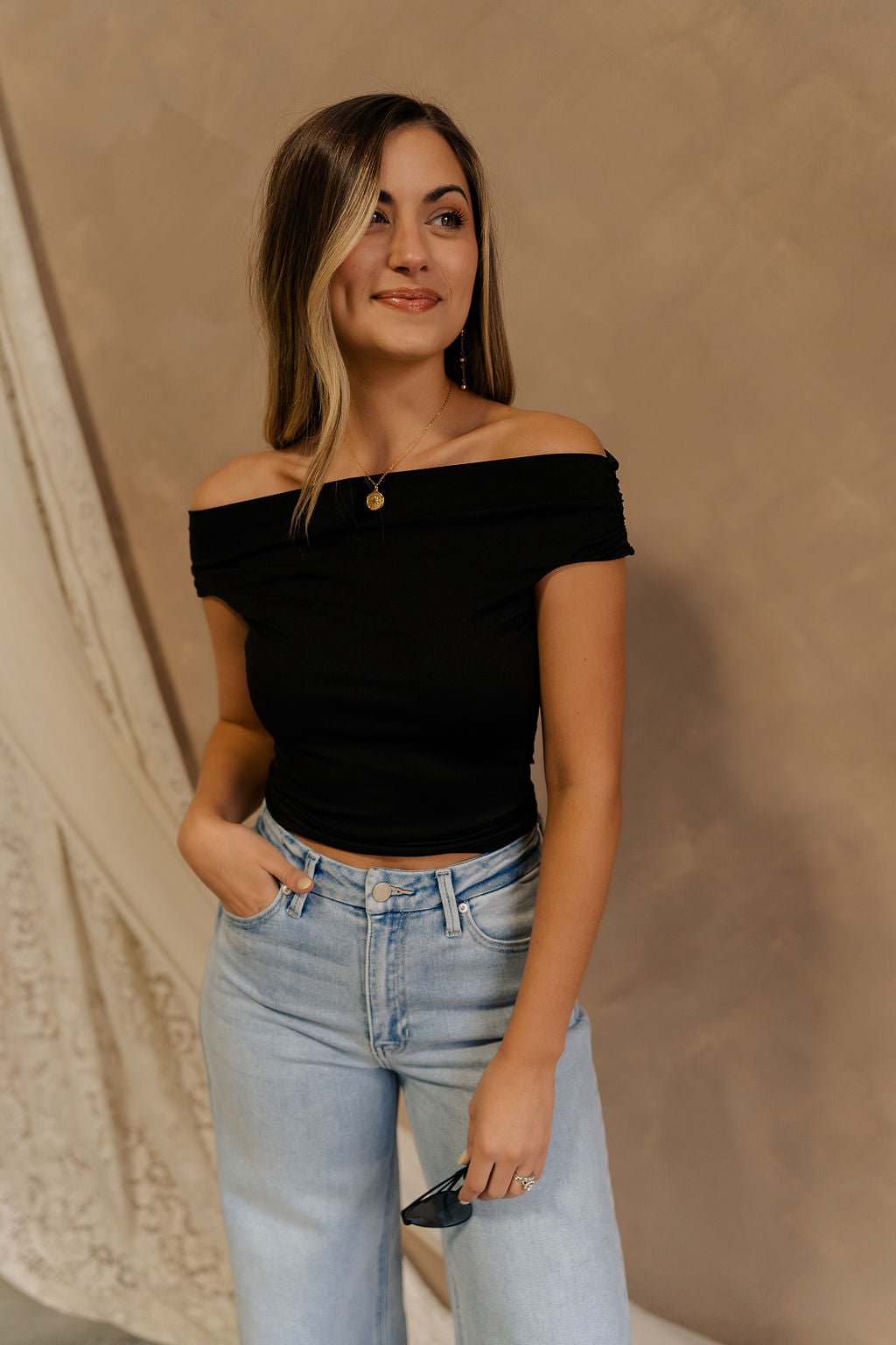 Front view of female model wearing the Isabella Off The Shoulder Cropped Tank in black, that features off the shoulder short sleeves, a straight neckline, and a cropped waist. Tank is paired with blue jeans.