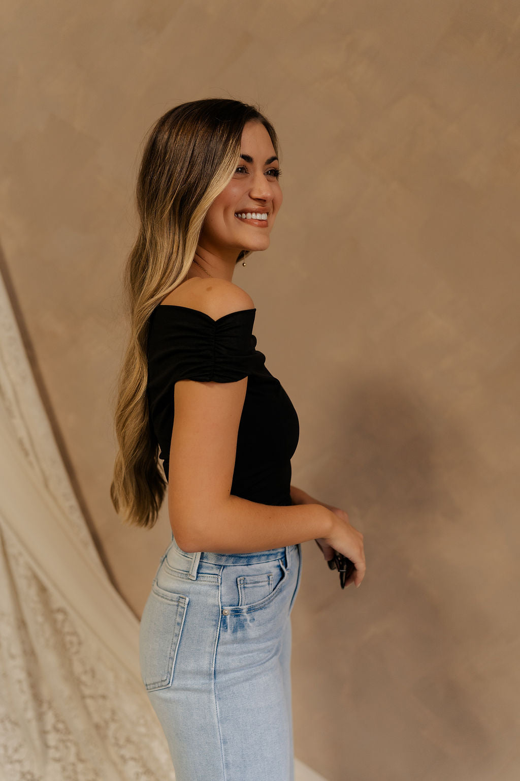 Side view of female model wearing the Isabella Off The Shoulder Cropped Tank in black, that features off the shoulder short sleeves, a straight neckline, and a cropped waist. Tank is paired with blue jeans.