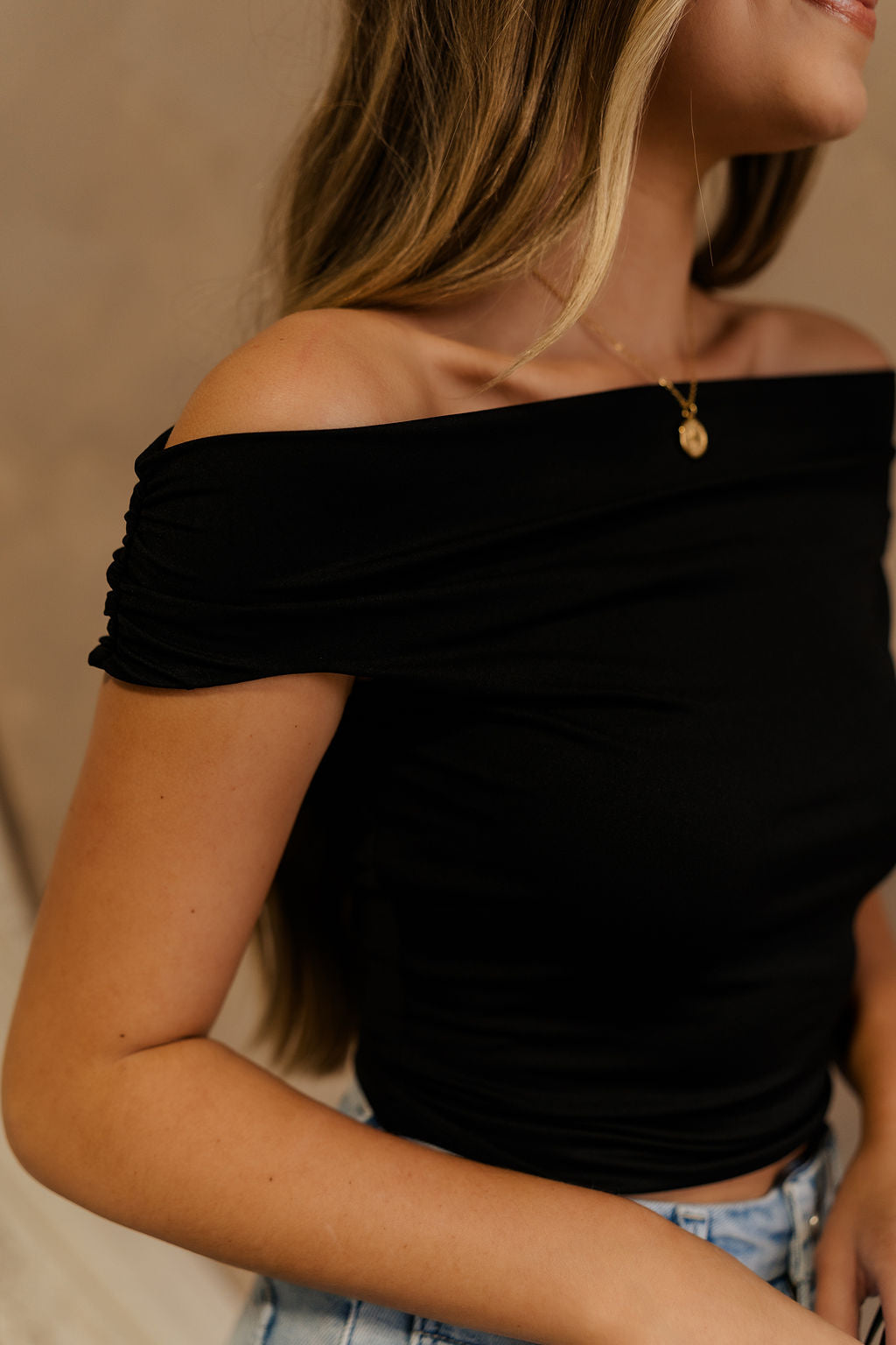 Close-up frontal side view of female model wearing the Isabella Off The Shoulder Cropped Tank in black, that features off the shoulder short sleeves, a straight neckline, and a cropped waist. 