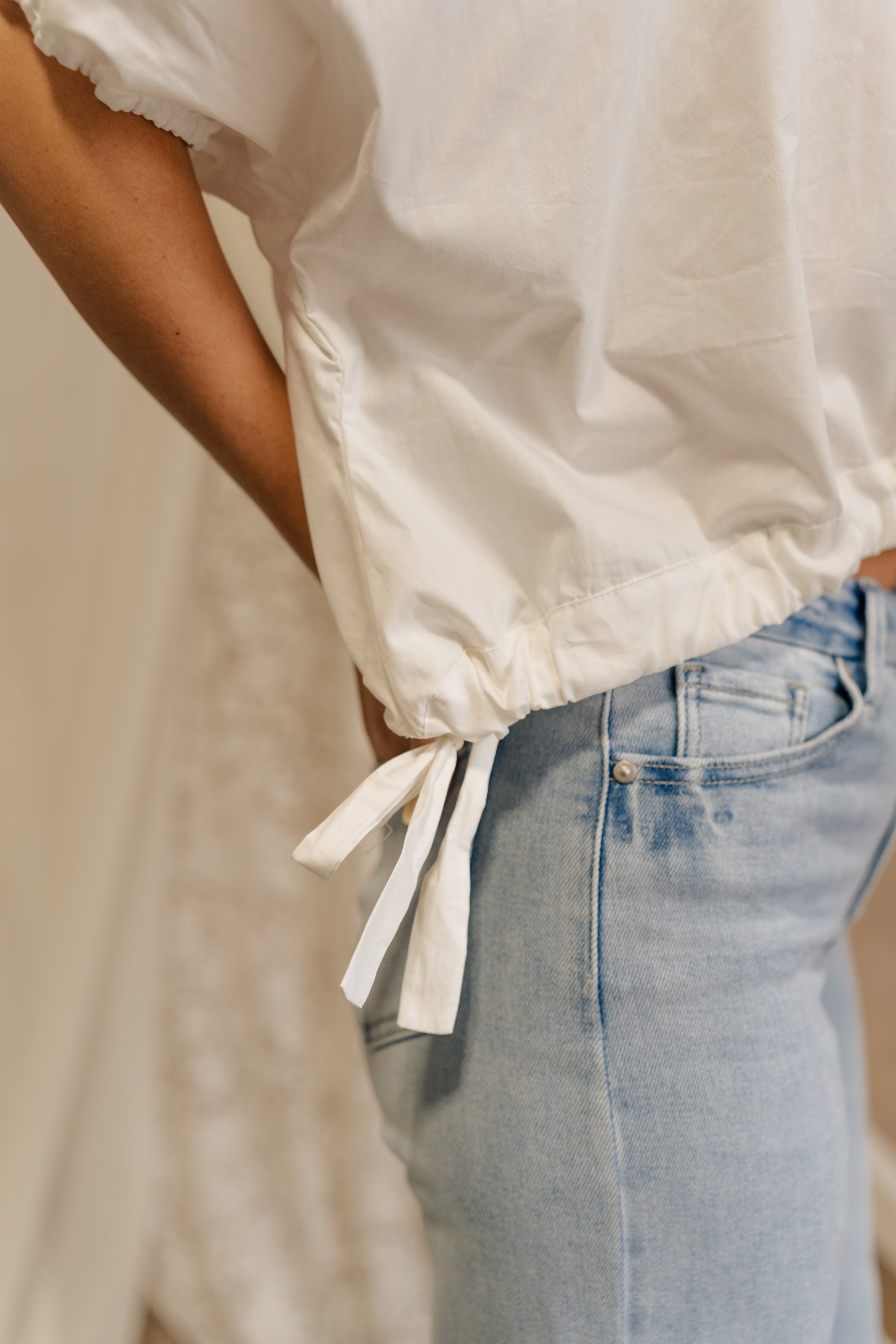 Close-up side view of female model wearing the Karina Drawstring Cropped Short Sleeve Top in white. This top has white fabric, short sleeves with elastic trim, and a cropped drawstring waist with ties.