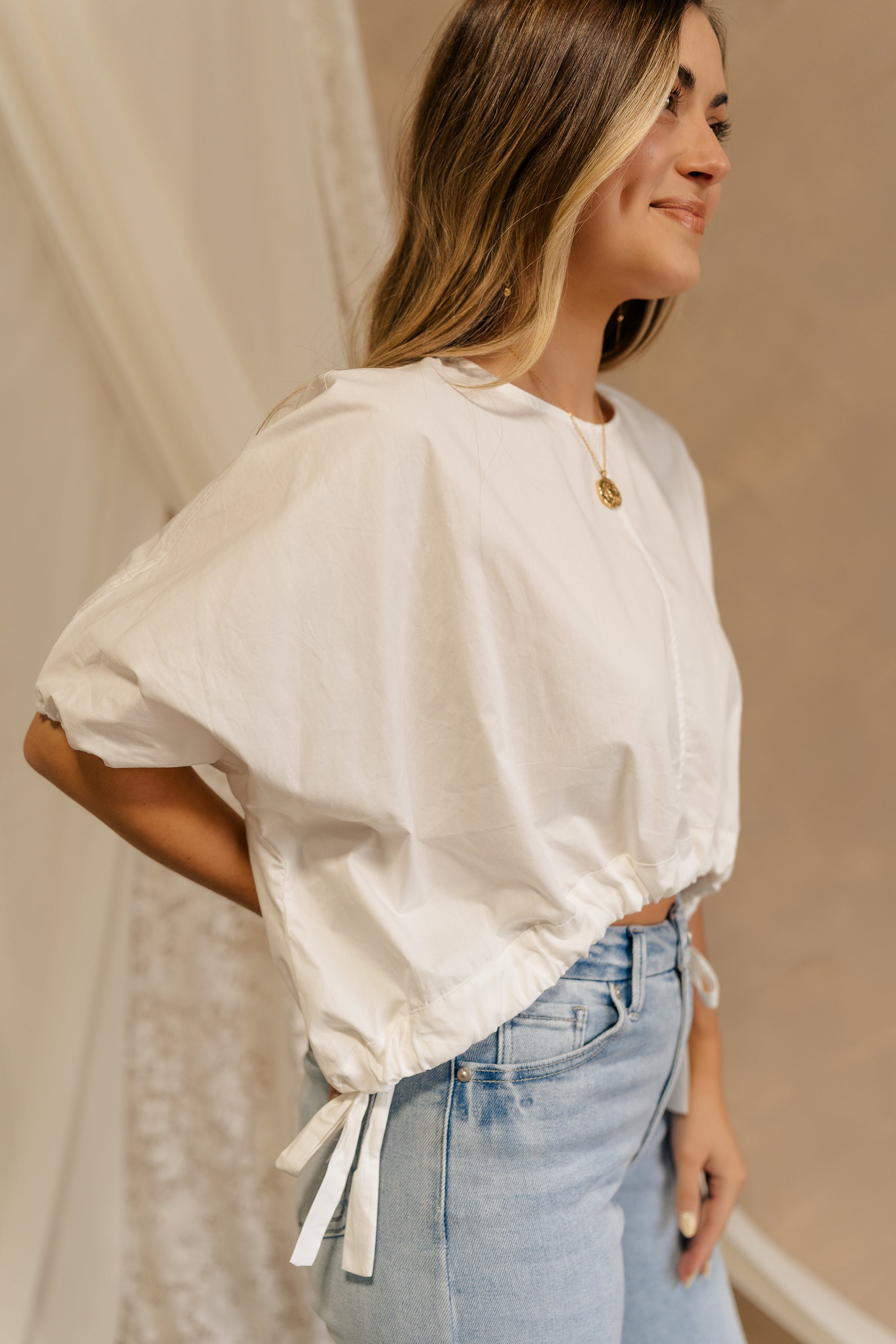 Side view of female model wearing the Karina Drawstring Cropped Short Sleeve Top in white. This top has white fabric, short sleeves with elastic trim, and a cropped drawstring waist with ties.