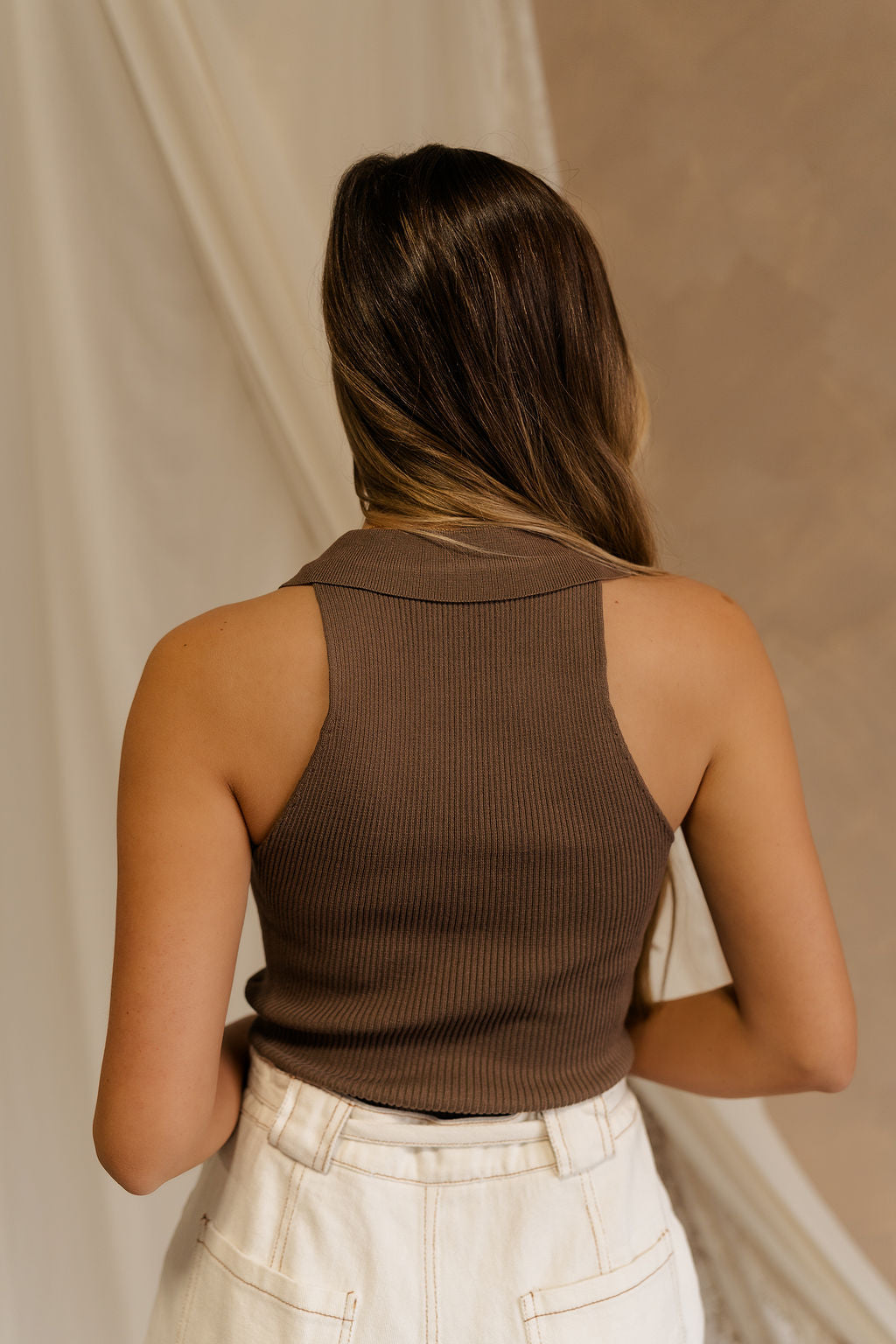 Back view of female model wearing the Maisie Ribbed Collar Tank in brown that feature brown ribbed fabric and a collared sleeveless neckline. Paired with white shotrs