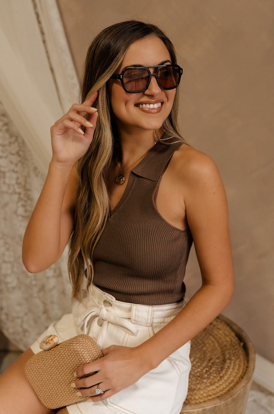 Side view of female model wearing the Maisie Ribbed Collar Tank in brown that feature brown ribbed fabric and a collared sleeveless neckline. Paired with white shotrs