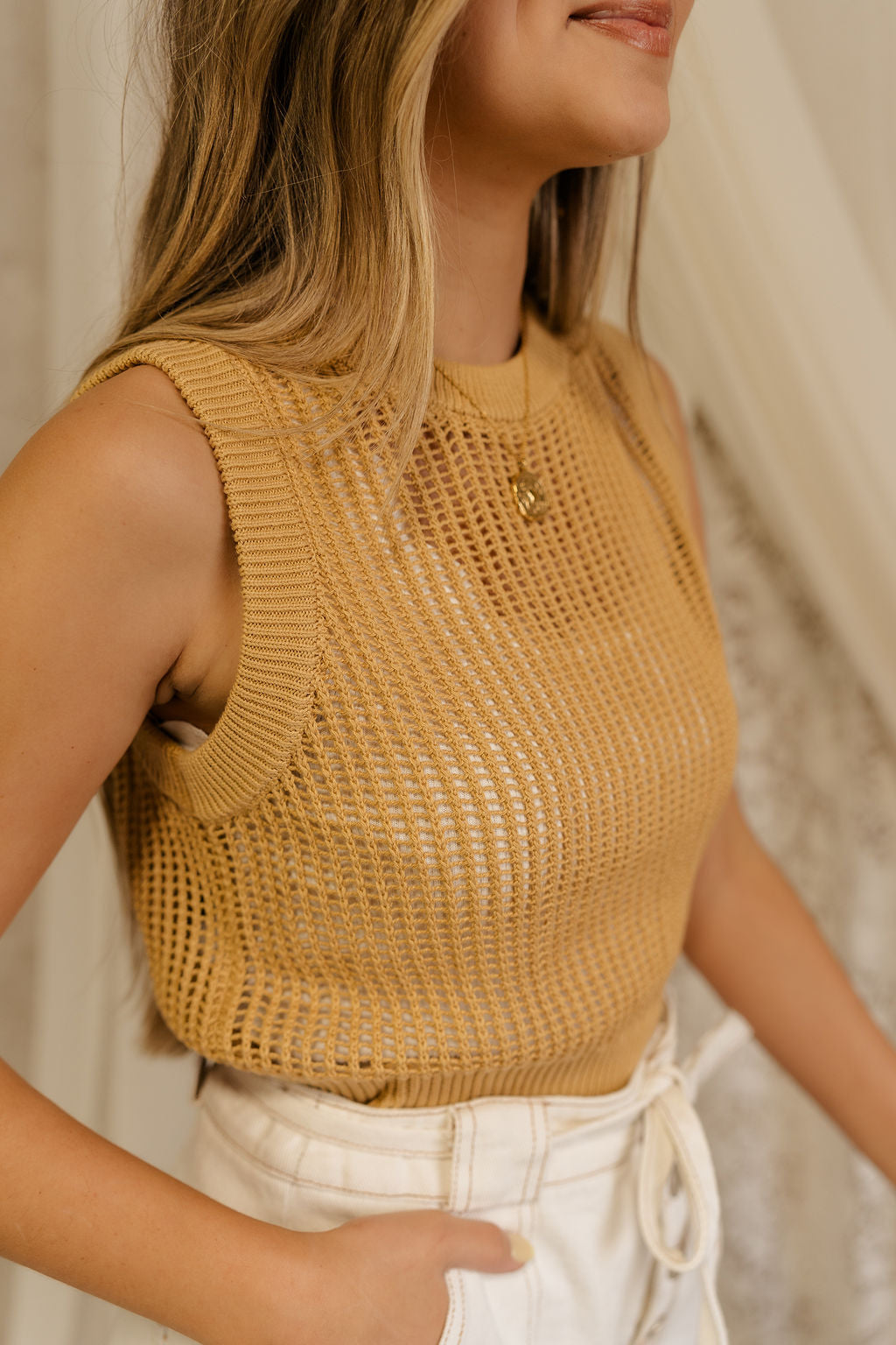Close side view of model wearing the Heidi Mustard Open Knit Tank which has a round neckline, open knit fabric, and sleeveless body. Layered over white tank top.