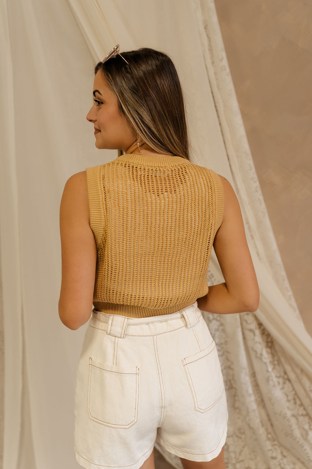 Back view of model wearing the Heidi Mustard Open Knit Tank which has a round neckline, open knit fabric, and sleeveless body. Layered over white tank top.