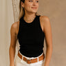 Front view of female model wearing the Winnie Black Ribbed Tank that has black ribbed fabric, sleeveless body, and round neck. Paired with brown belt and white jeans.