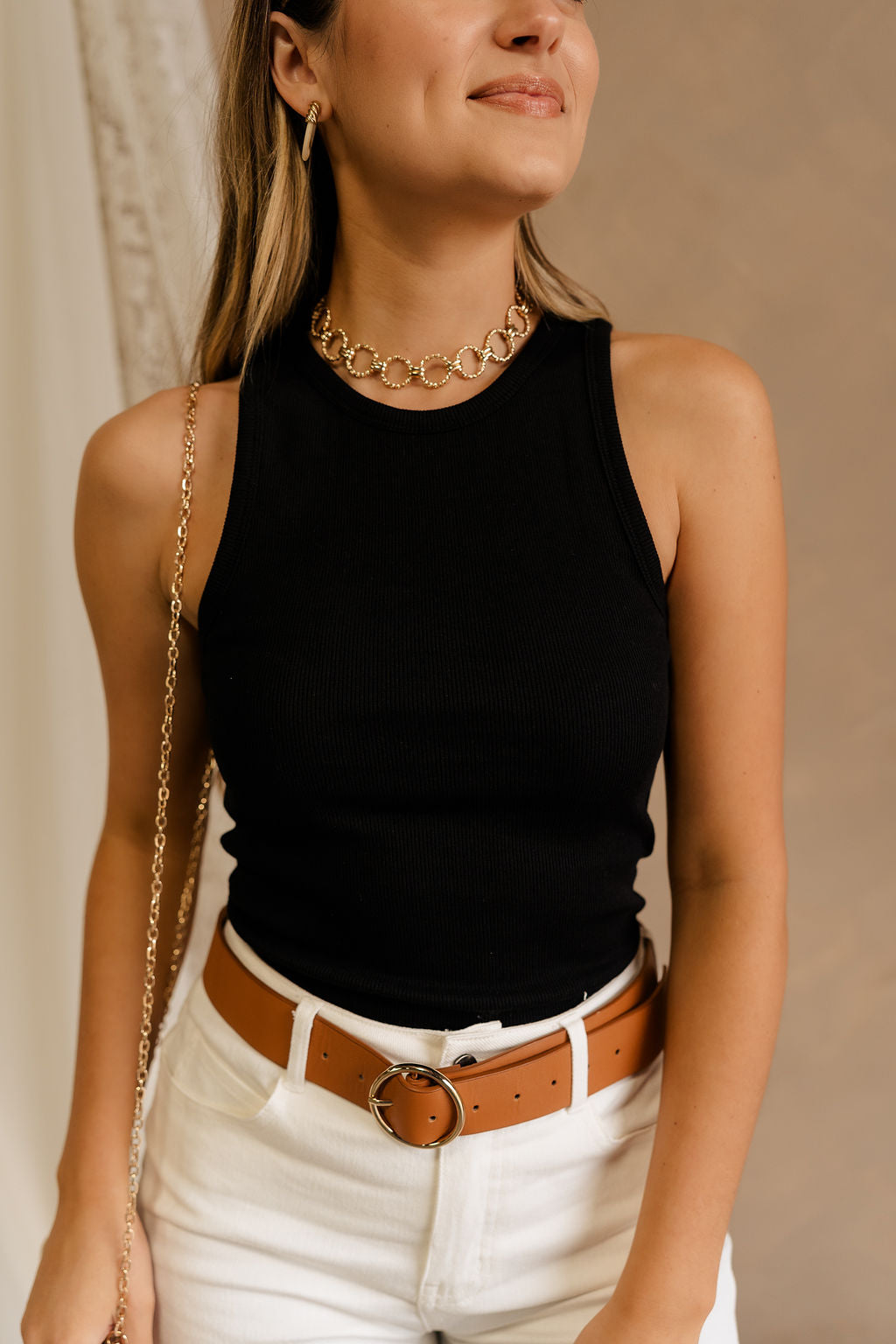 Close front view of female model wearing the Winnie Black Ribbed Tank that has black ribbed fabric, sleeveless body, and round neck. Paired with brown belt and white jeans.