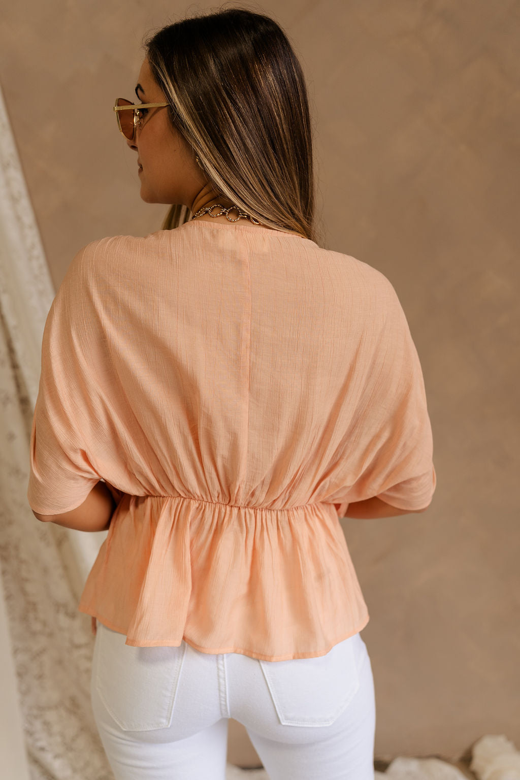 Back view of female model wearing the Juliet Peach Ruched Short Sleeve Top that features flowy peach fabric, ruched short sleeves, and an elastic waist.