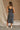 Full body back view of female model wearing the Leyla Navy & White Striped Midi Dress that has knit fabric, navy trim, a side slit, thick straps, and a square neck.
