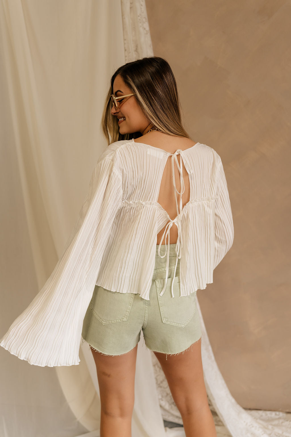 Back view of female model wearing the Kimberly Cream Pleated Long Sleeve Top which features Cream Sheer Plisse Fabric, Cropped Waist, Lettuce Hem Details, Round Neckline and Open Back with Tie Closures