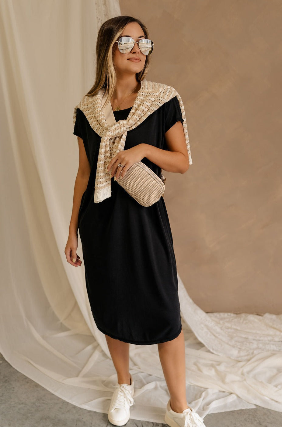 Full body front view of female model wearing the Remy Black Short Sleeve Midi Dress that features black knit fabric, short sleeves, midi length, and side slits. Worn with Rebecca Ivory Sling Bag and sweater around her shoulders.