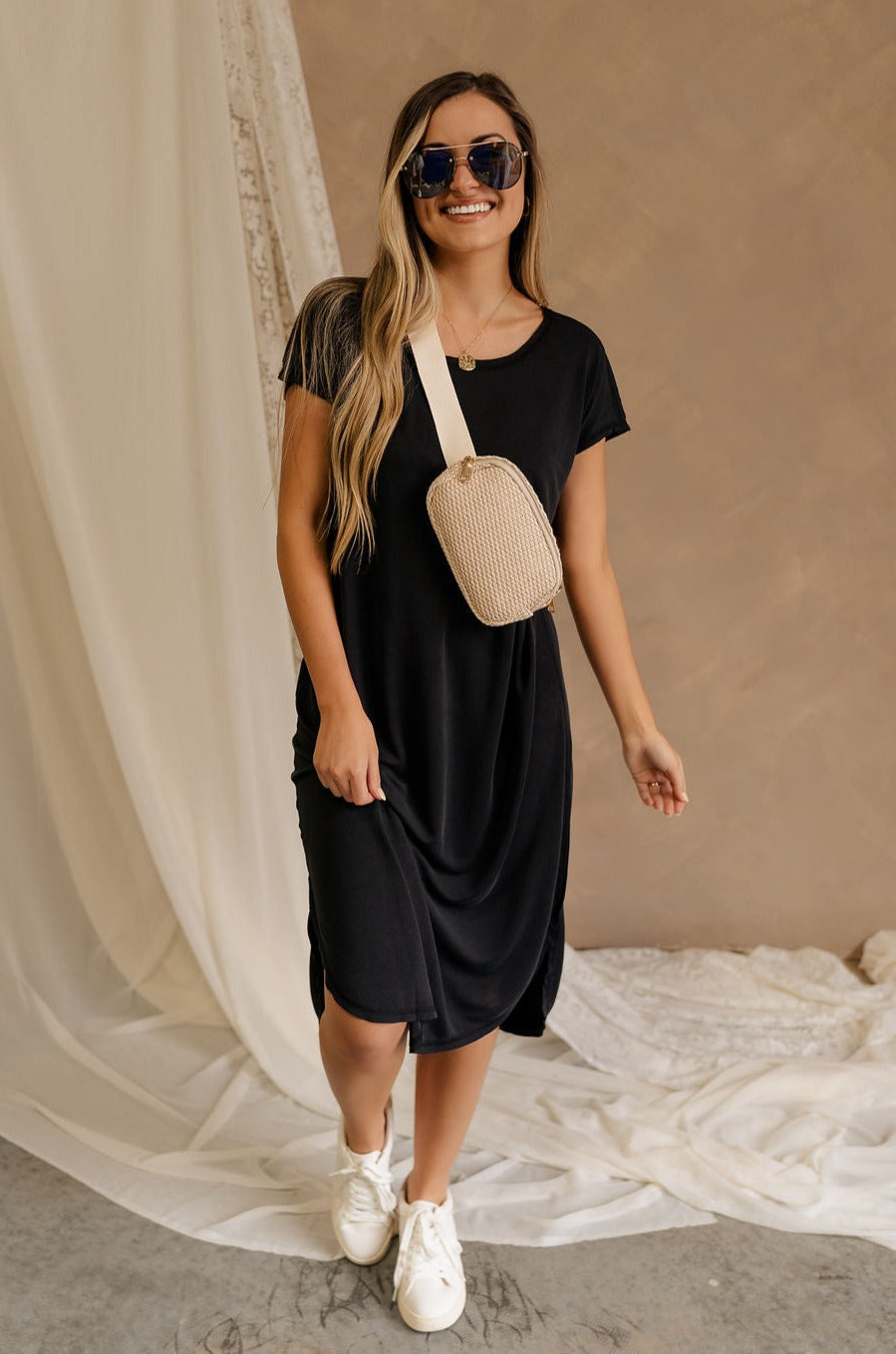 Full body front view of female model wearing the Remy Black Short Sleeve Midi Dress that features black knit fabric, short sleeves, midi length, and side slits. Worn with Rebecca Ivory Sling Bag.