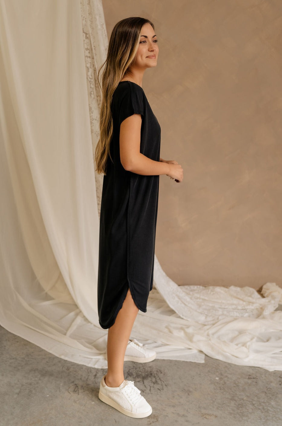 Full body side view of female model wearing the Remy Black Short Sleeve Midi Dress that features black knit fabric, short sleeves, midi length, and side slits.