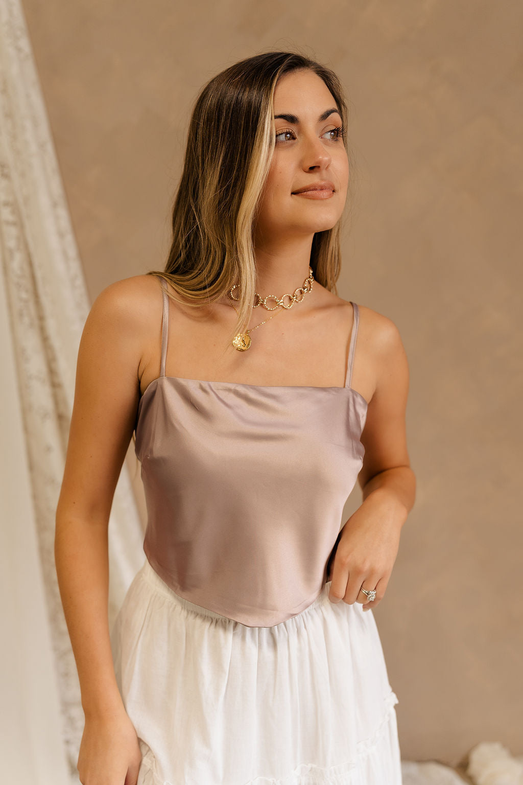 Front view of female model wearing the Jovie Mauve Satin Tank that has mauve satin fabric, thin straps, and a tie back. Paired with white skirt.