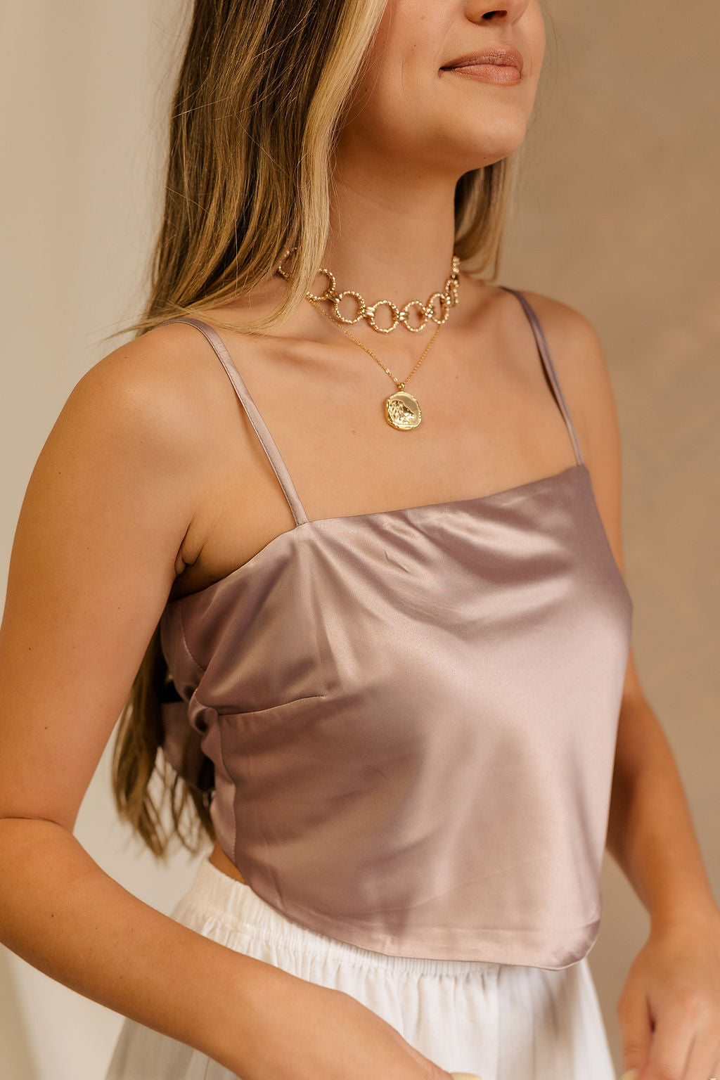 Close side view of female model wearing the Jovie Mauve Satin Tank that has mauve satin fabric, thin straps, and a tie back. Paired with white skirt.