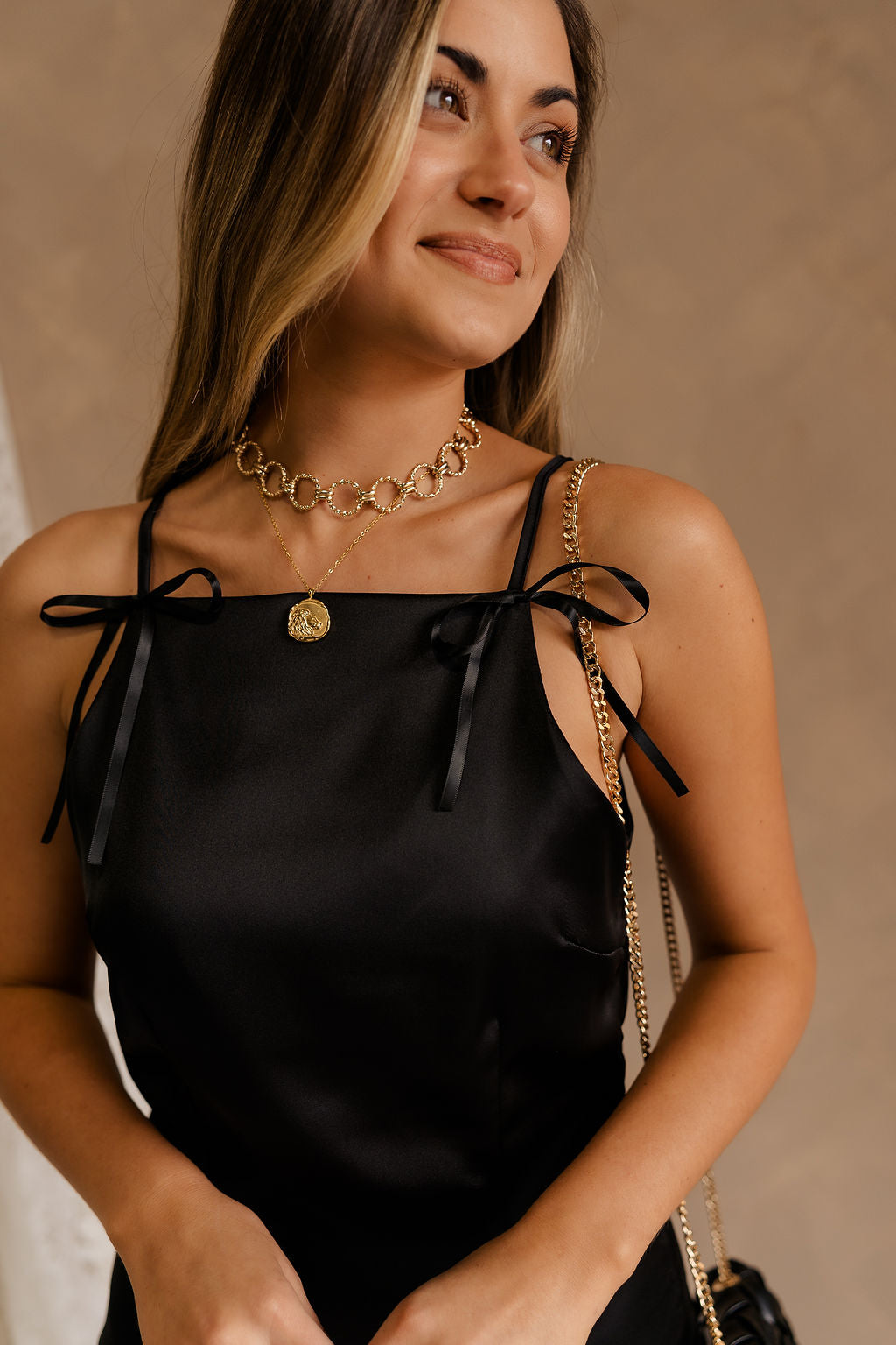 Upper body front view of model wearing the Monica Black Satin Mini Dress that features black satin fabric, thin straps with bow details, a square neck, and mini length.