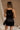 Back view of model wearing the Monica Black Satin Mini Dress that features black satin fabric, thin straps with bow details, a square neck, and mini length.