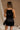 Back view of model wearing the Monica Black Satin Mini Dress that features black satin fabric, thin straps with bow details, a square neck, and mini length.