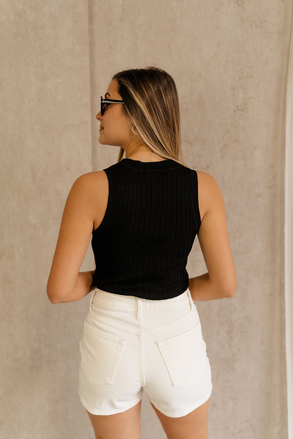 Back view of model wearing the Rayna Black Ribbed Tank, that features wide ribbed fabric, a round neckline, and sleeveless body. Worn with white shotrs.