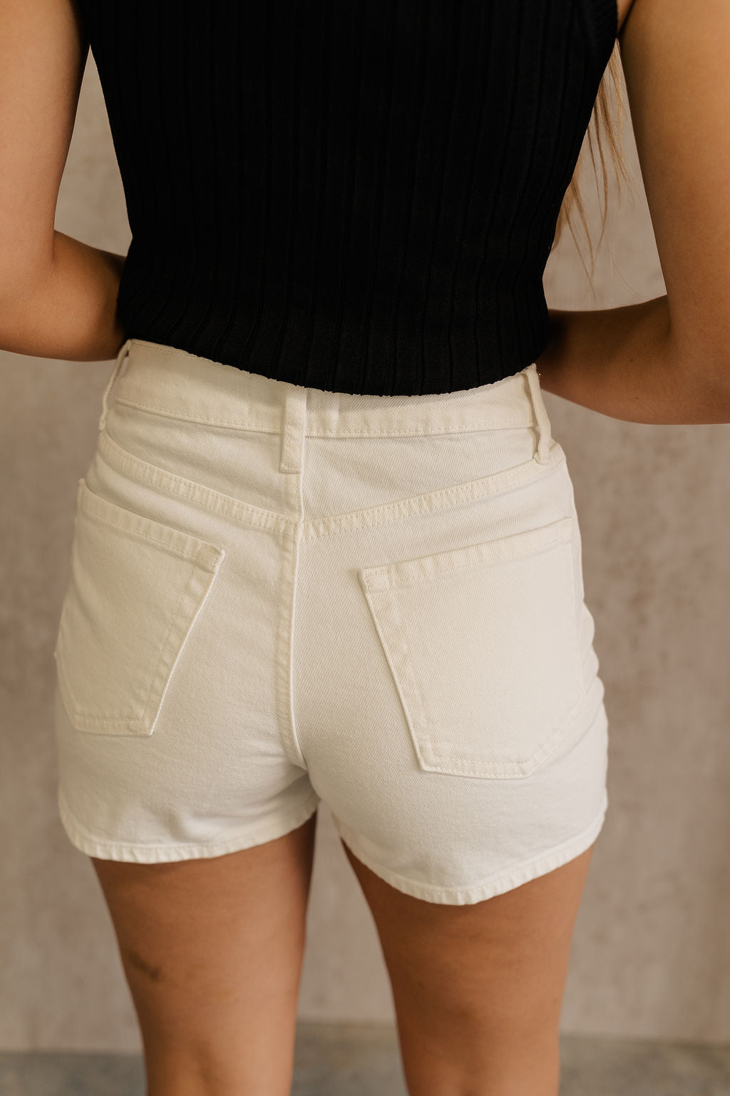 Back view of female model wearing the Andi White Denim Shorts that have white denim fabric, a button fly with silver buttons, belt loops, and front and back pockets. Worn with black tank.