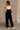 Full body back view of female model wearing the Jessie Wide Leg Jumpsuit in Black, that features black woven material, adjustable straps, and wide legs. Worn over white tank top.